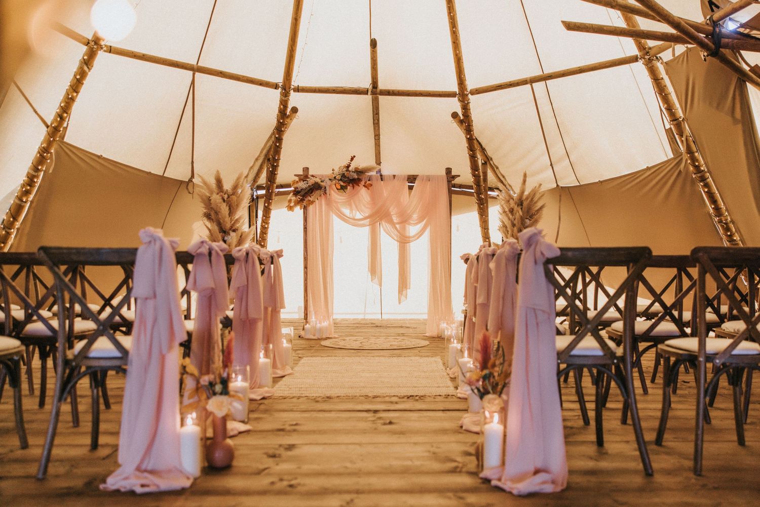 Interior of a rustic tipi wedding venue with wooden aisle, cross-back chairs and flowing fabric drapes in warm lighting at Nuddock Wood Lake