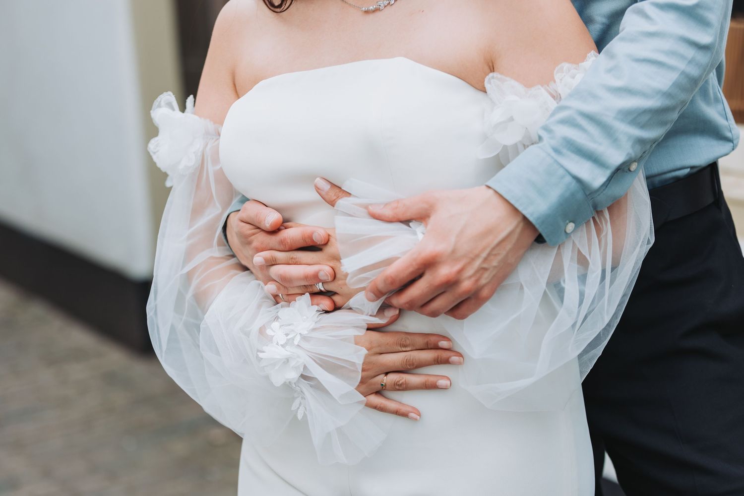 Close up of hands embracing around a white wedding dress with delicate fabric details.