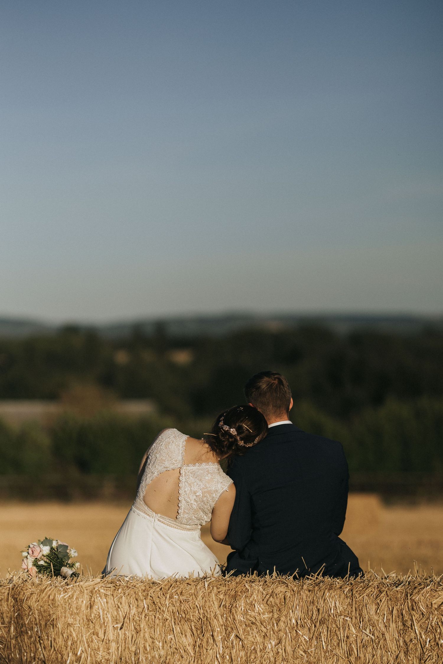 Newlywed couple sits together watching sunset on hilltop overlooking rural landscape with golden field.