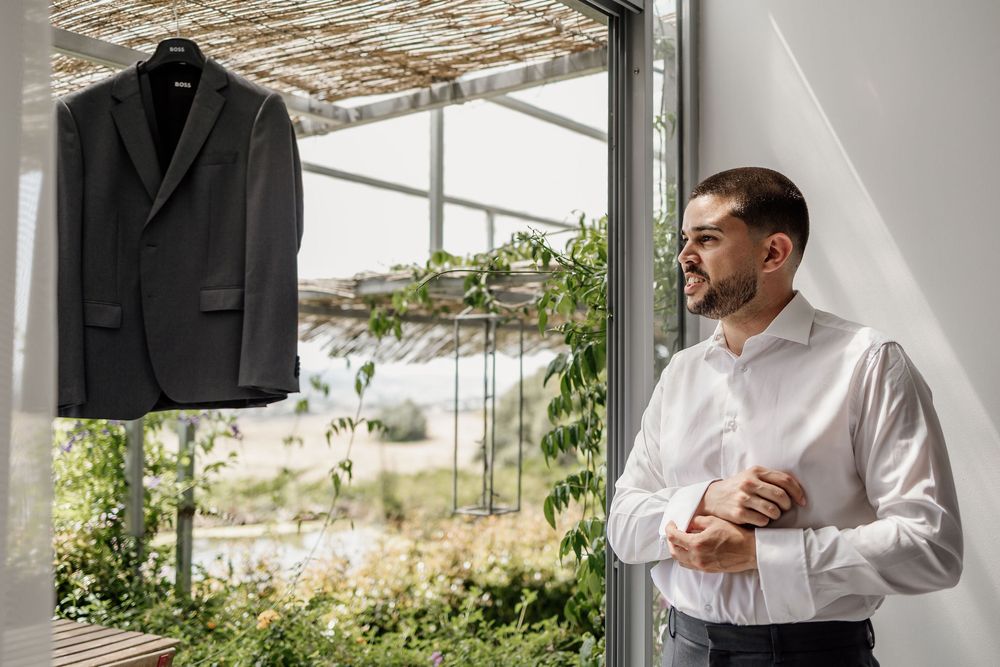 A dark suit jacket hangs while someone in a white dress shirt stands by a window in a sunlit room with plants.