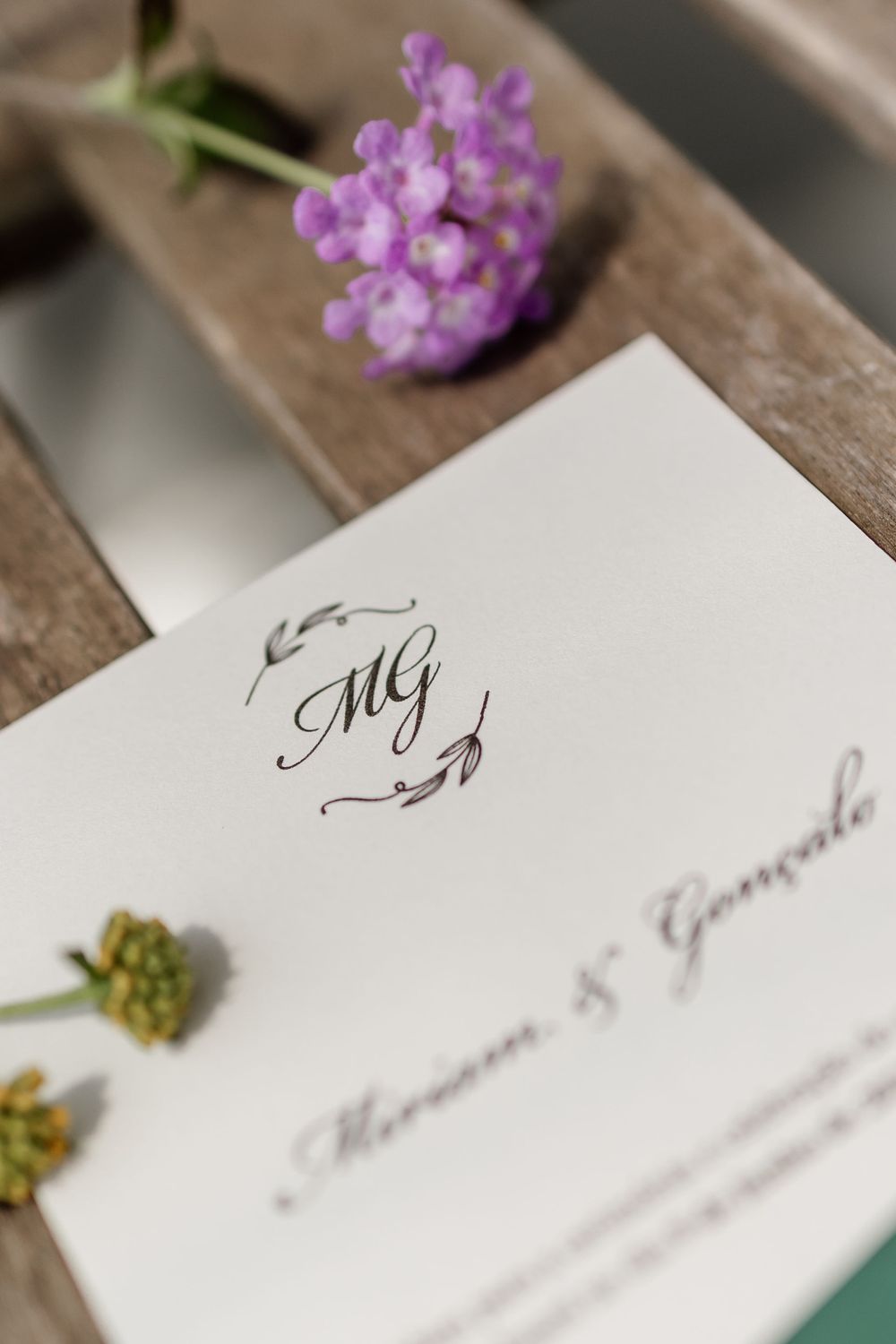 Wedding invitation with elegant monogram and purple flowers on rustic wooden surface in soft lighting.