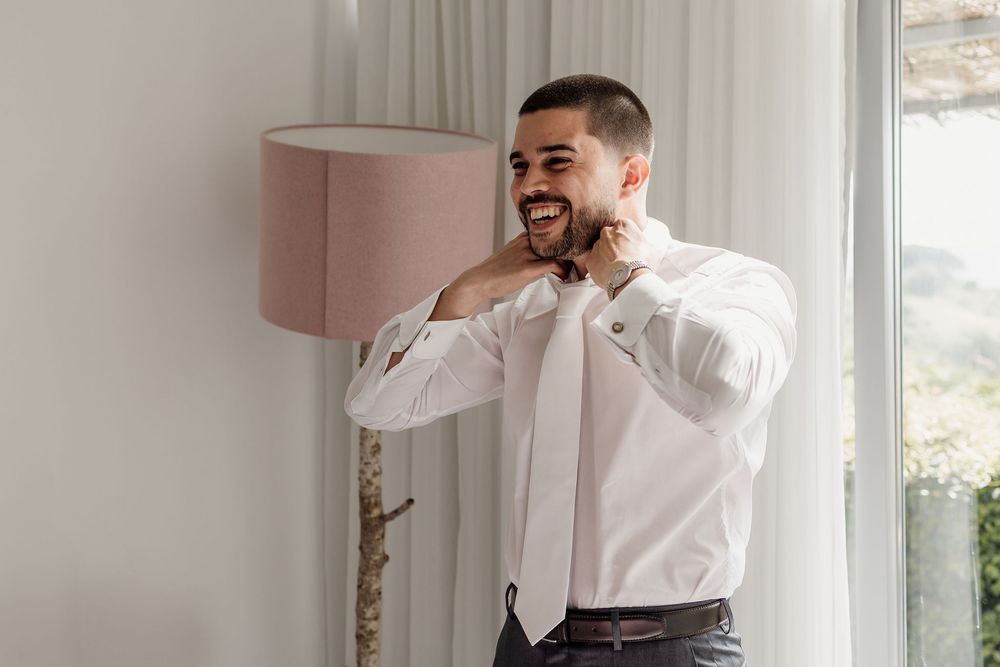 Someone adjusts their white tie while preparing in a light-filled room with a pink lampshade and window.