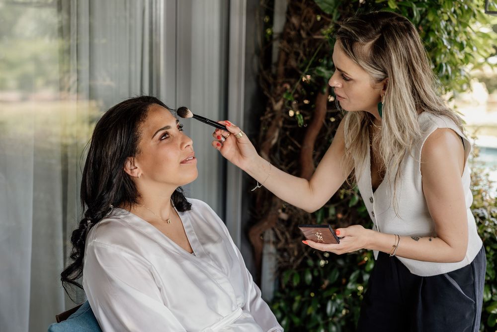 A makeup artist applies cosmetics to a client getting ready while sitting in front of glass doors.