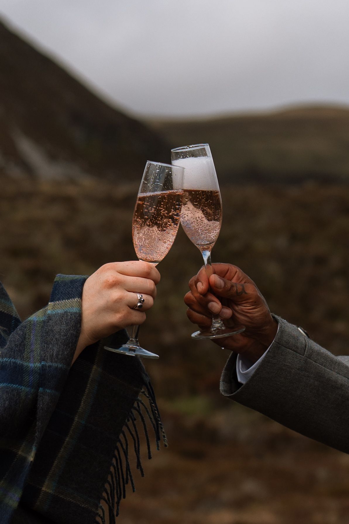 Two people toast with glasses of sparkling rose champagne against a moody mountain landscape background.
