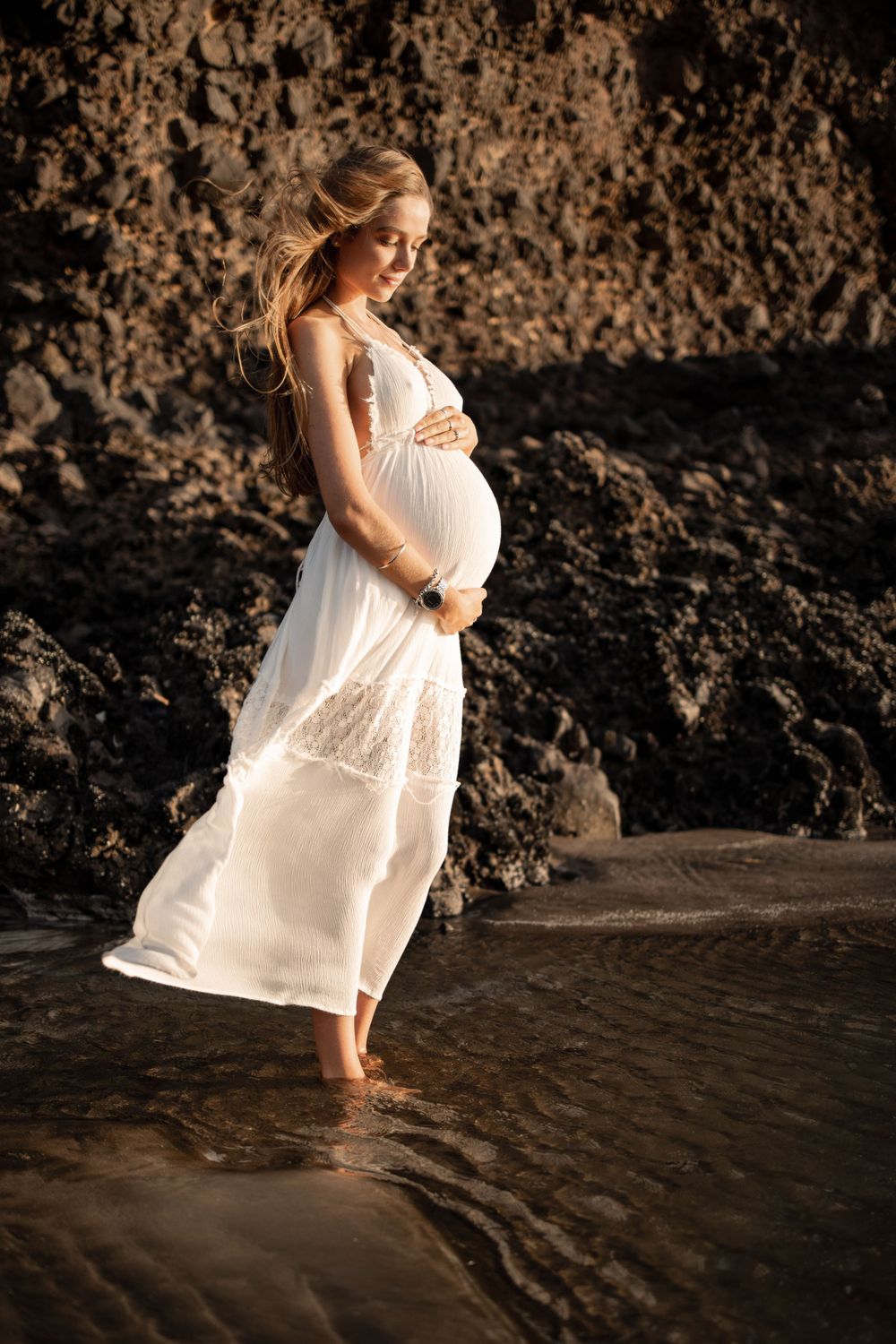 Pregnant woman stands in water cradling her belly at a beach for a golden hour maternity photoshoot. Rocky background
