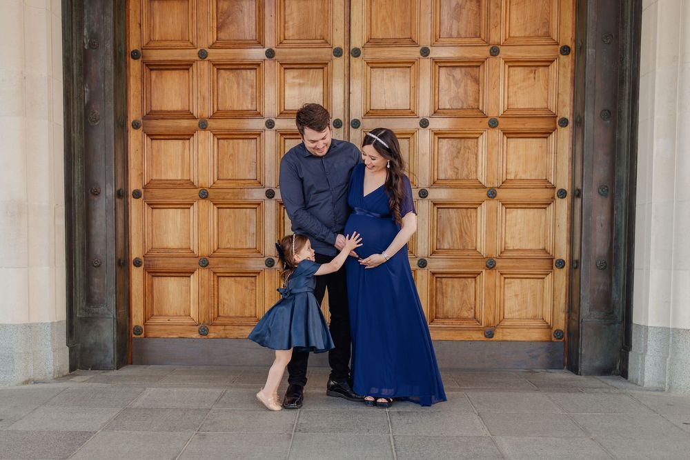 Family embracing outside the main entrance of Auckland museum. Daughter holds her hand up to Mother's pregnant belly. Father embraces them