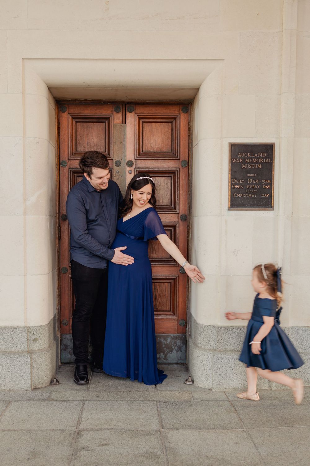 Girls runs to her heavily pregnant mother's arms in front of beautiful wooden door. Outside Auckland museum. Father embraces mother smiling