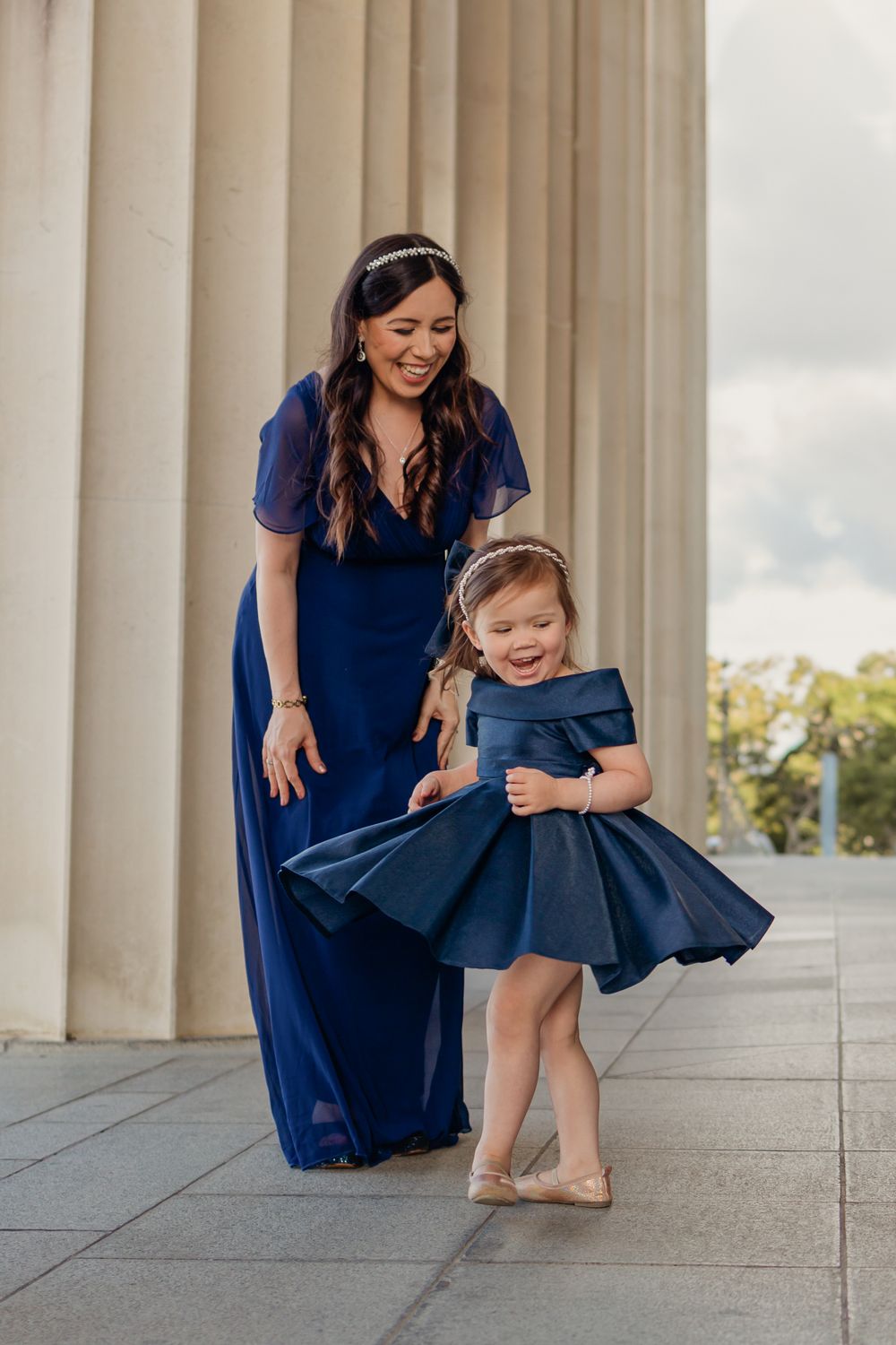 Little girl twirls around with her dress swirling. Her mother stands behind her smiling in awe of her. Large pillar in the background.