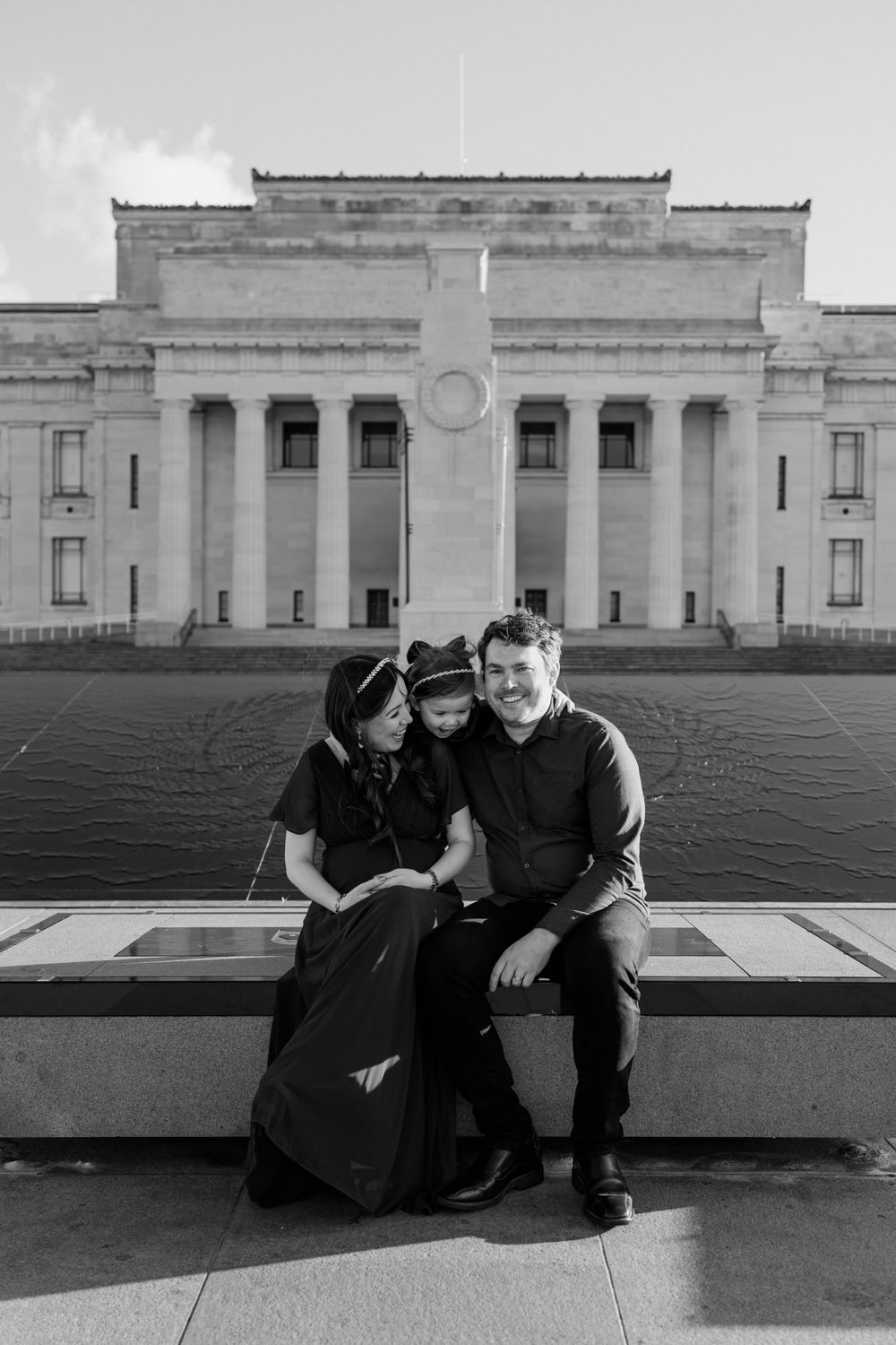 Black and white family portrait of pregnant Mother, Father and daughter sitting by the fountain with the Auckland Museum in the background.