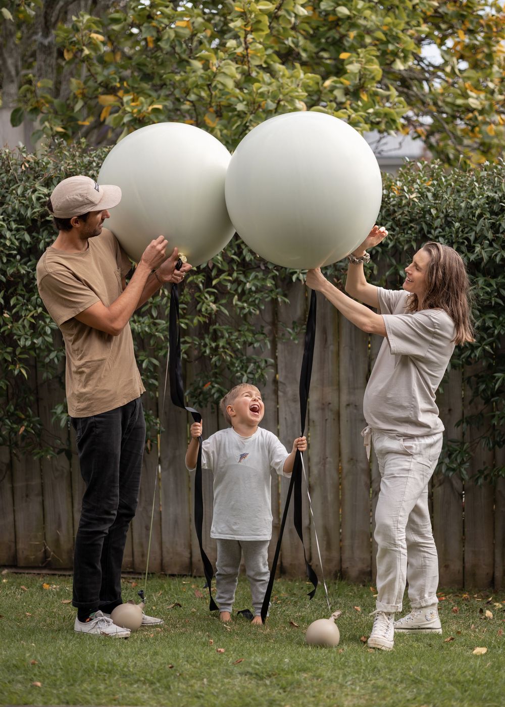 Mother and Father hold pin to balloons filled with coloured confetti outdoors for gender reveal as son runs up to parents.