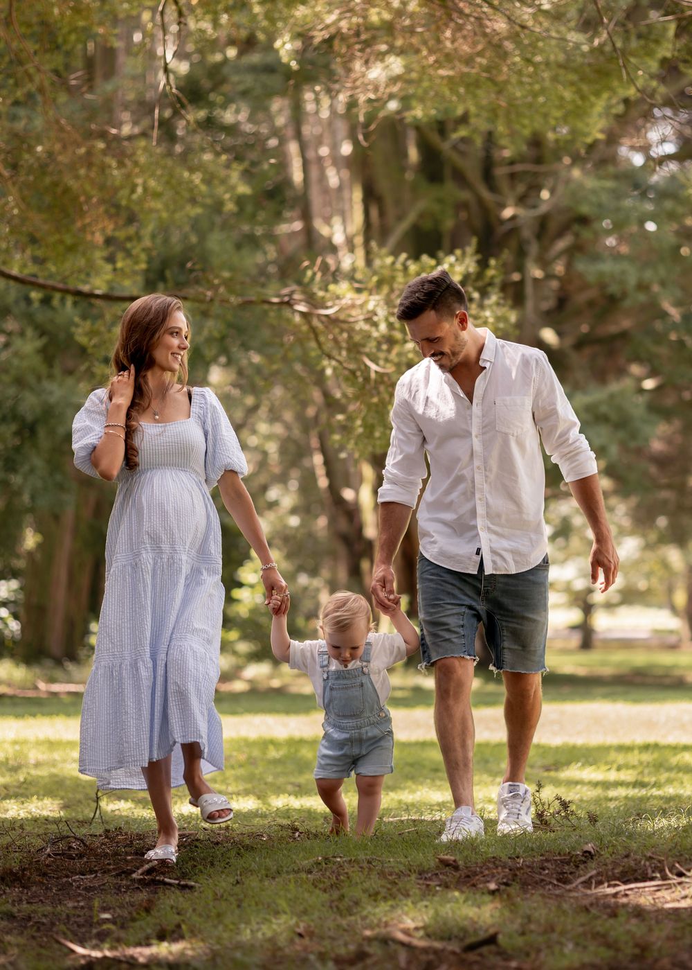 Mother, father and toddler holding hands as they walk towards the camera in a forrest setting for a maternity photoshoot