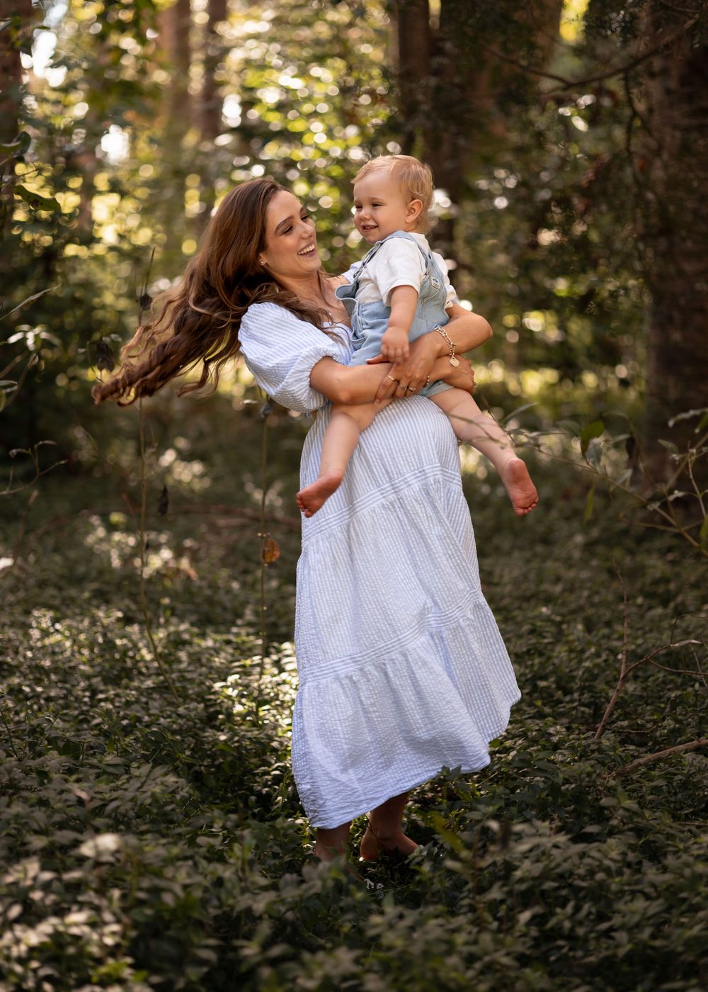 Pregnant mother holding toddler as they swing around. Deep in the forest, golden light shining through leaves. Maternity Photoshoot