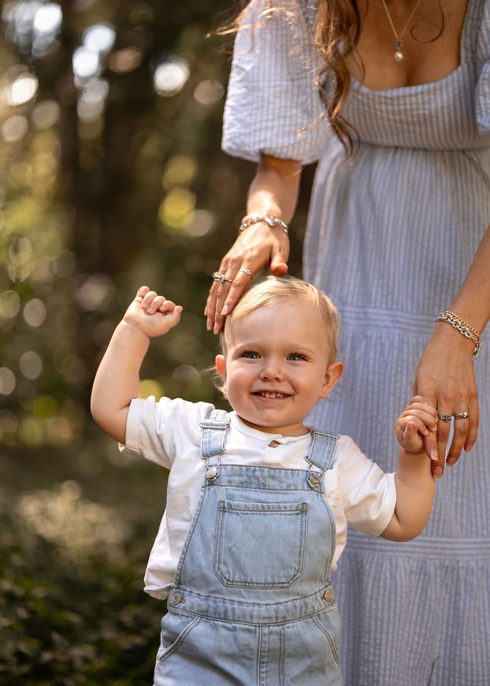 Smiling toddler wearing denim overalls holds his mother's hand as she lovingly pats his head in a forest setting