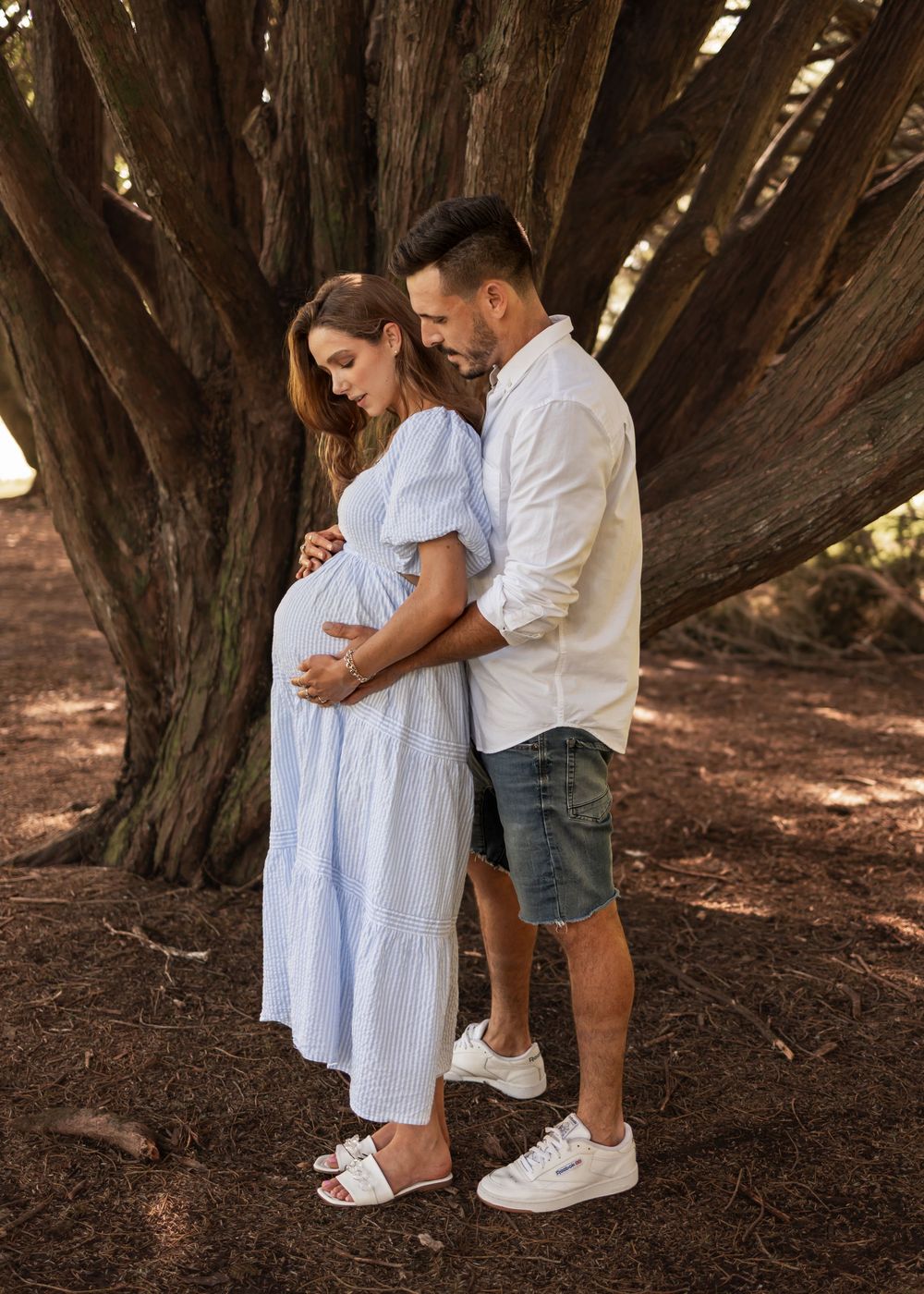 Husband embraces pregnant wife from behind in front of a large tree. Beautiful earthy tones in a forest setting. looking at her baby bump