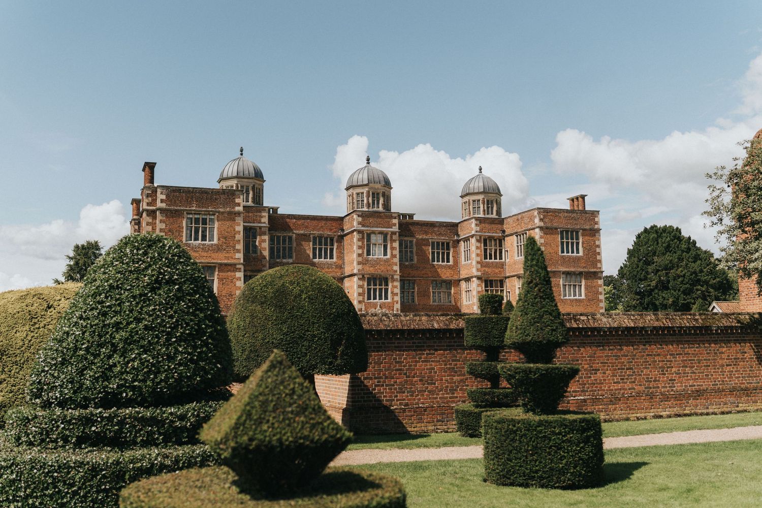 Historic red brick Tudor mansion with ornate chimneys and decorative topiary gardens featuring sculpted evergreen bushes.
