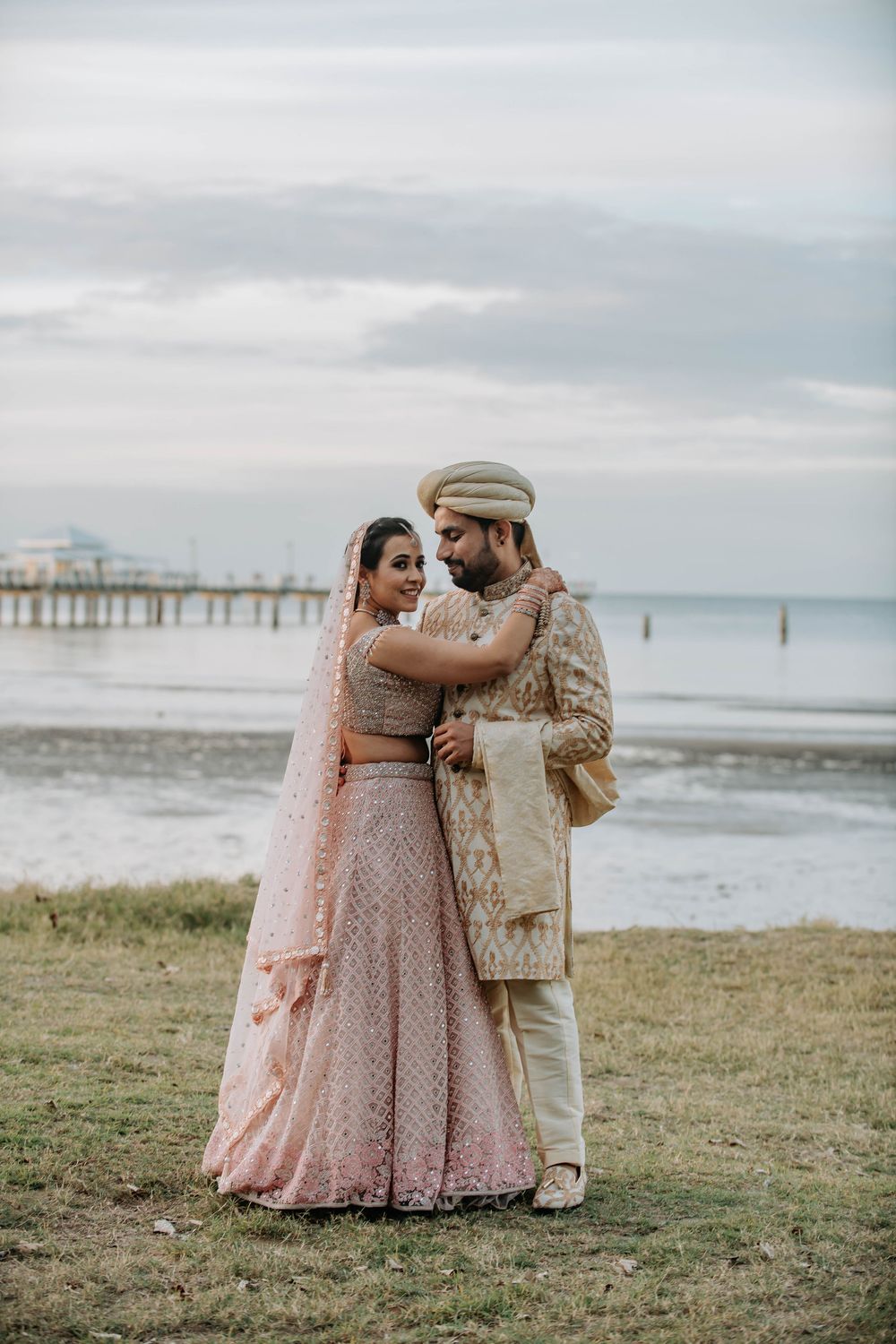 A couple in traditional Indian wedding attire embrace while standing on grass near a pier at the beach during sunset.
