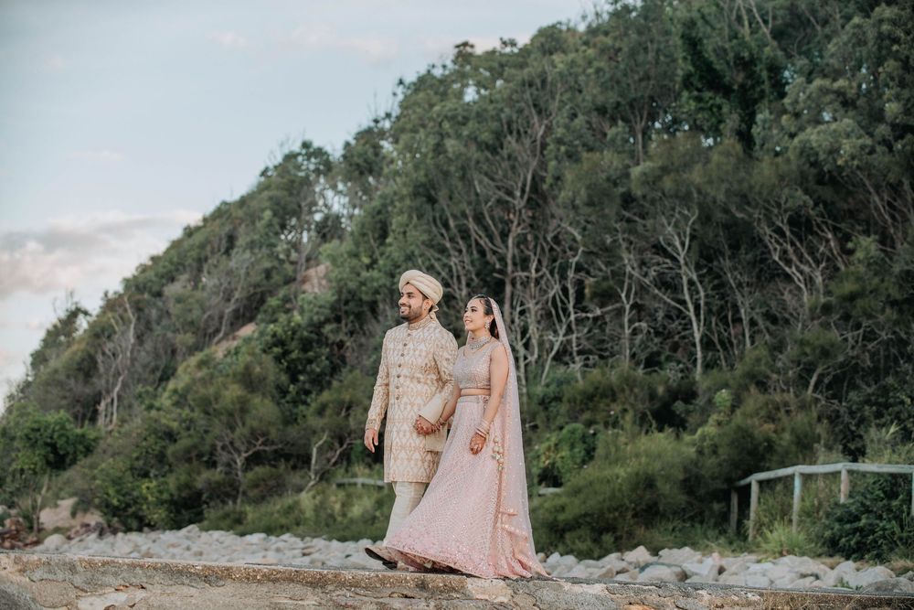 Two people in formal attire walk together on a rocky beach path with lush green forest in the background at sunset.