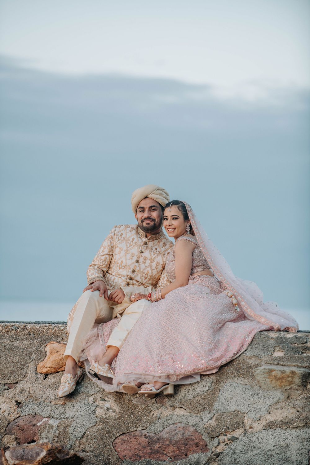 A joyful couple in traditional South Asian wedding attire sit together on a stone wall against a cloudy sky.