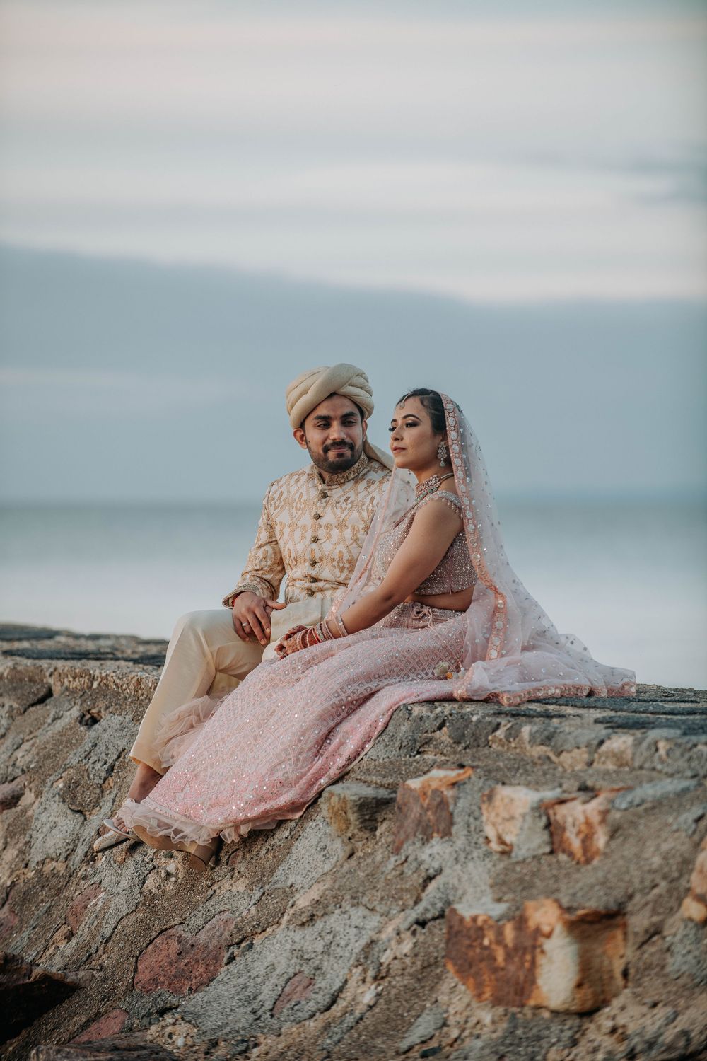 A romantic couple in traditional South Asian wedding attire sits together on a stone wall at sunset.