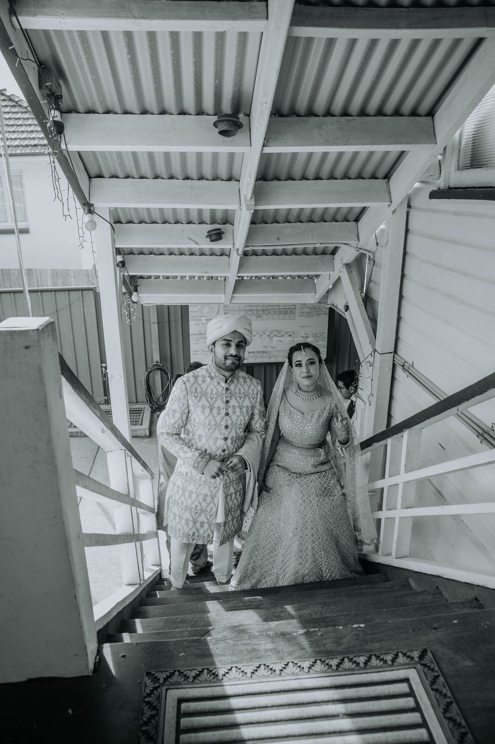 Black and white photo of a couple in traditional Indian wedding attire descending stairs under a covered walkway.