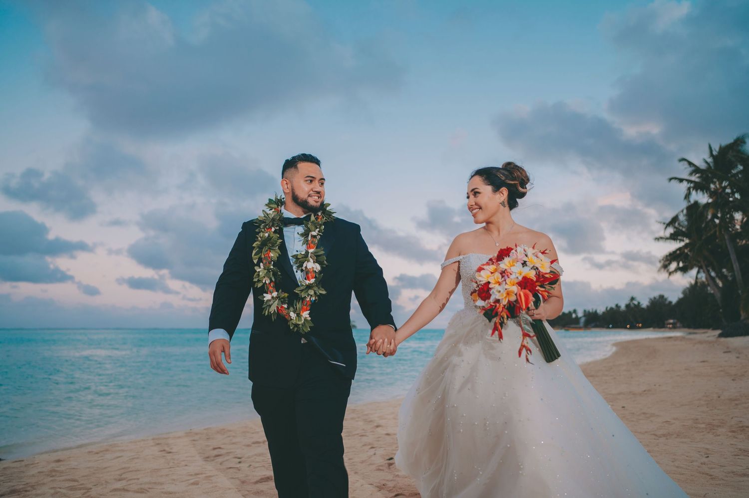 Wedding couple poses against scenic beach sunset with palm trees and tropical backdrop.