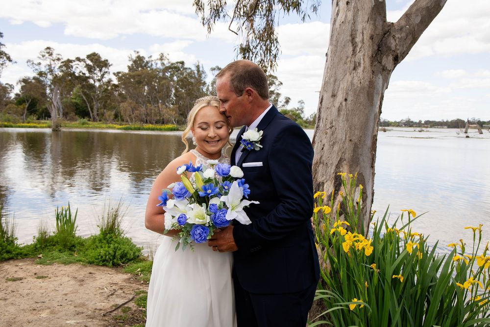 A romantic wedding couple embraces by a serene lake with blooming yellow irises and eucalyptus trees in the background.