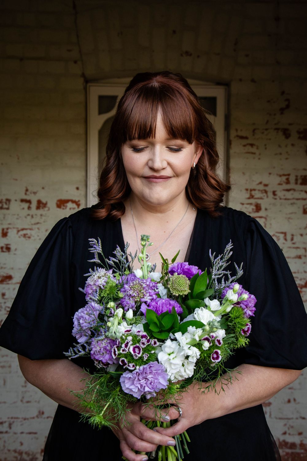 Purple and white floral bouquet arrangement being held against a rustic brick wall.