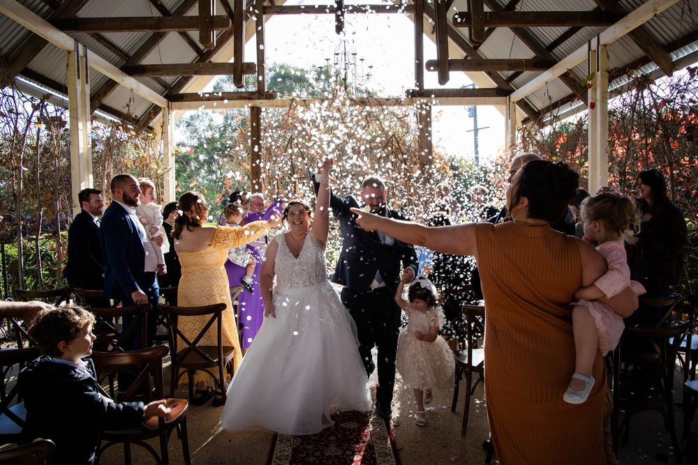 Wedding celebration with guests showering confetti over the newlyweds in a rustic indoor venue with exposed beams.