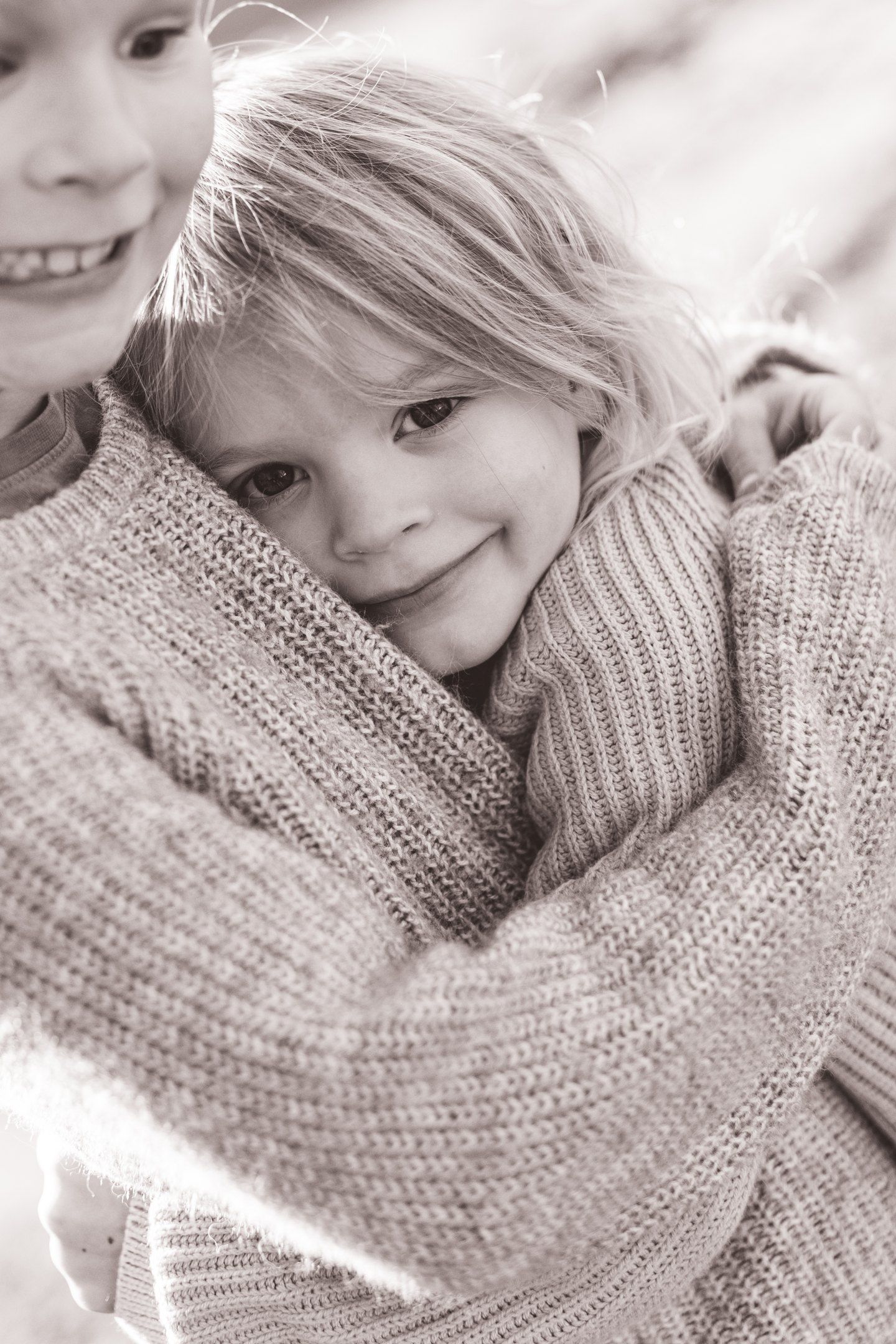 Two young children in cozy knit sweaters share a warm embrace outdoors in a charming black and white photo.