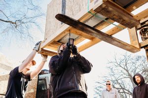 Group of people at a construction site with wooden beams overhead looking upward against a bright blue sky.