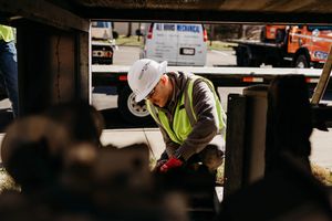 Construction worker in safety vest and hard hat performs maintenance work underneath equipment at job site.
