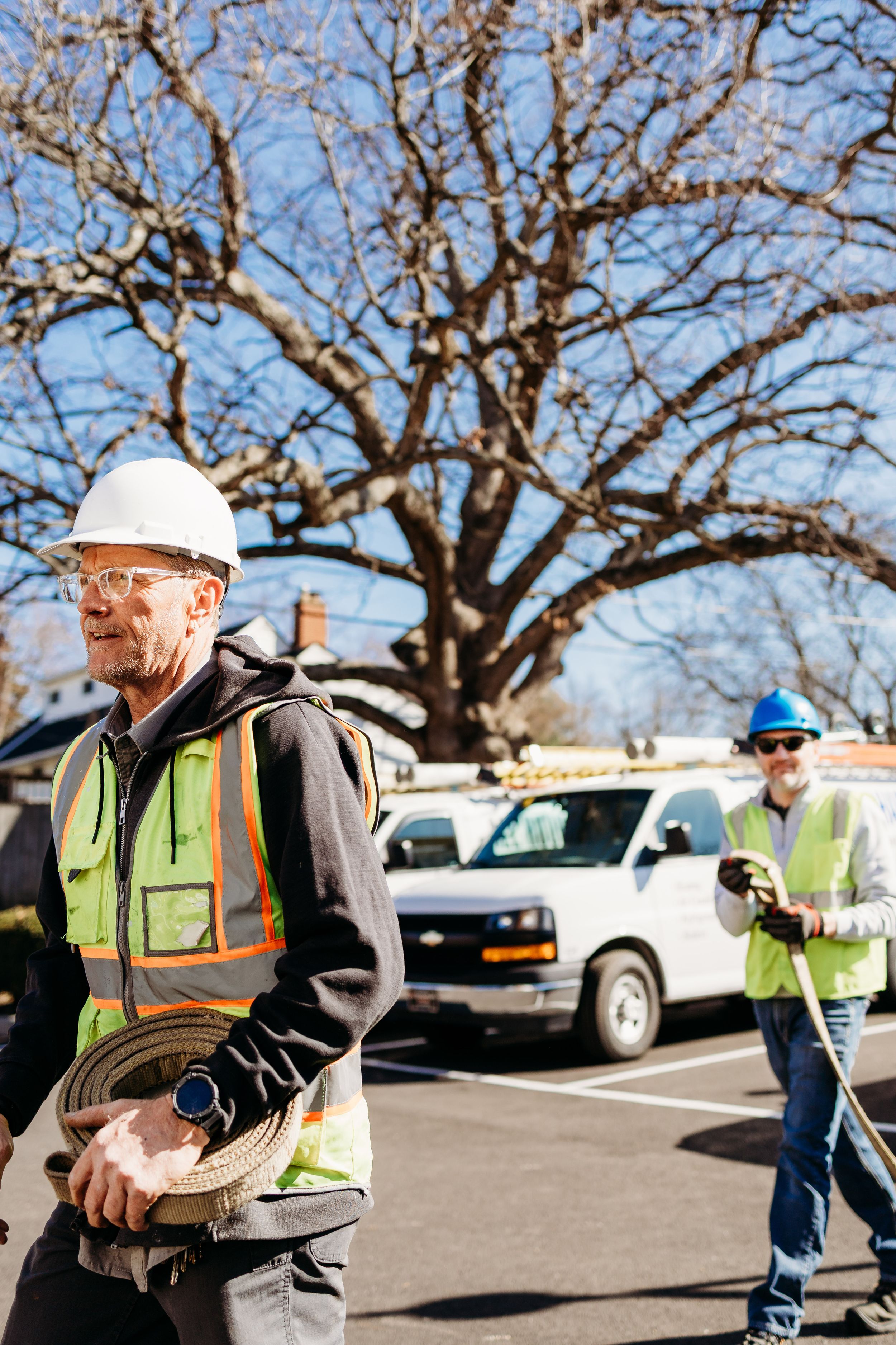 Construction worker in safety vest and hard hat carries equipment across parking lot on sunny day.