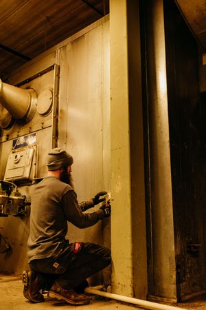 Contractor in protective gear performing pipe maintenance tasks in dimly lit basement corridor.