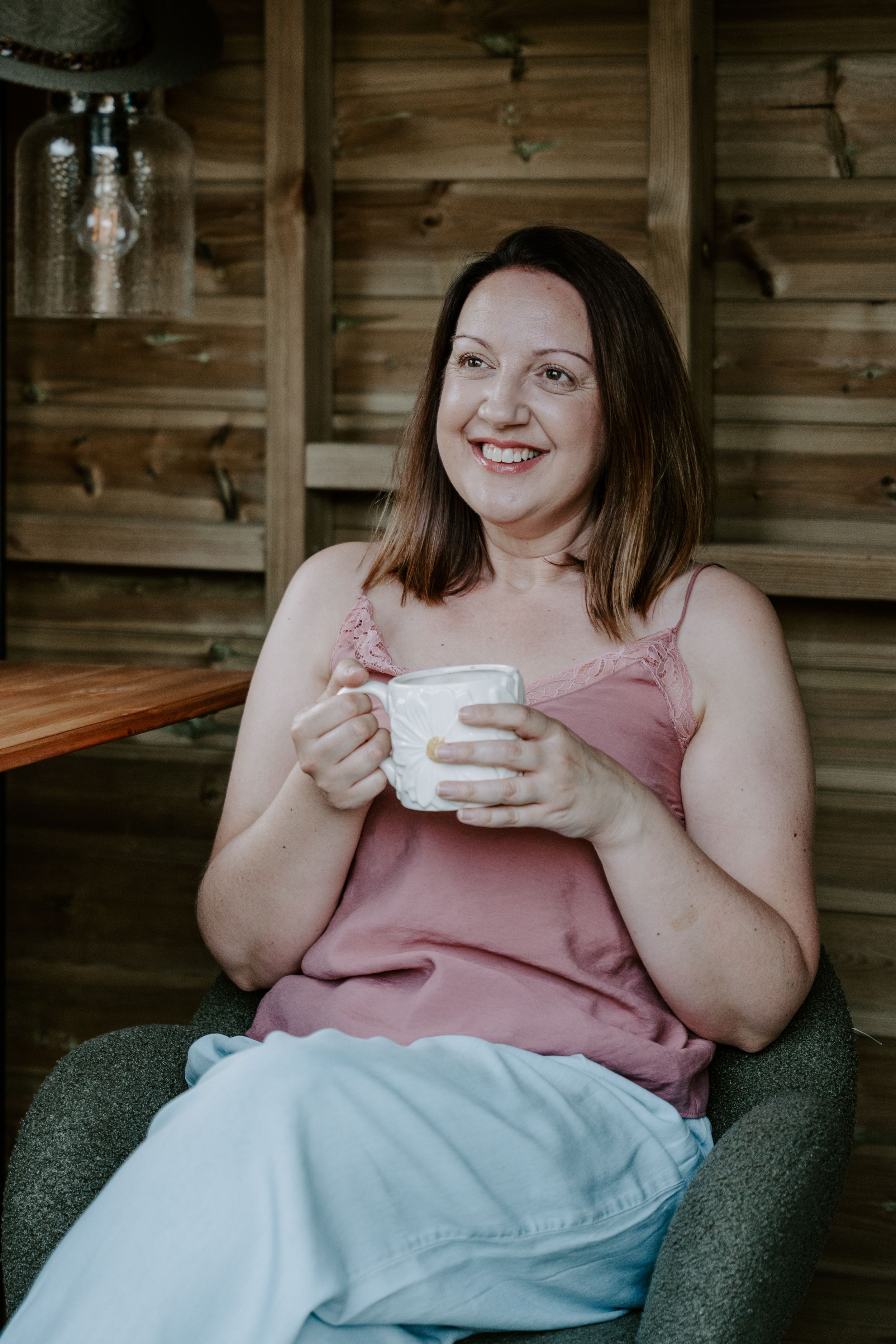 A photo of a lady sitting in a summer house holding a cup of tea and smiling away from the camera.