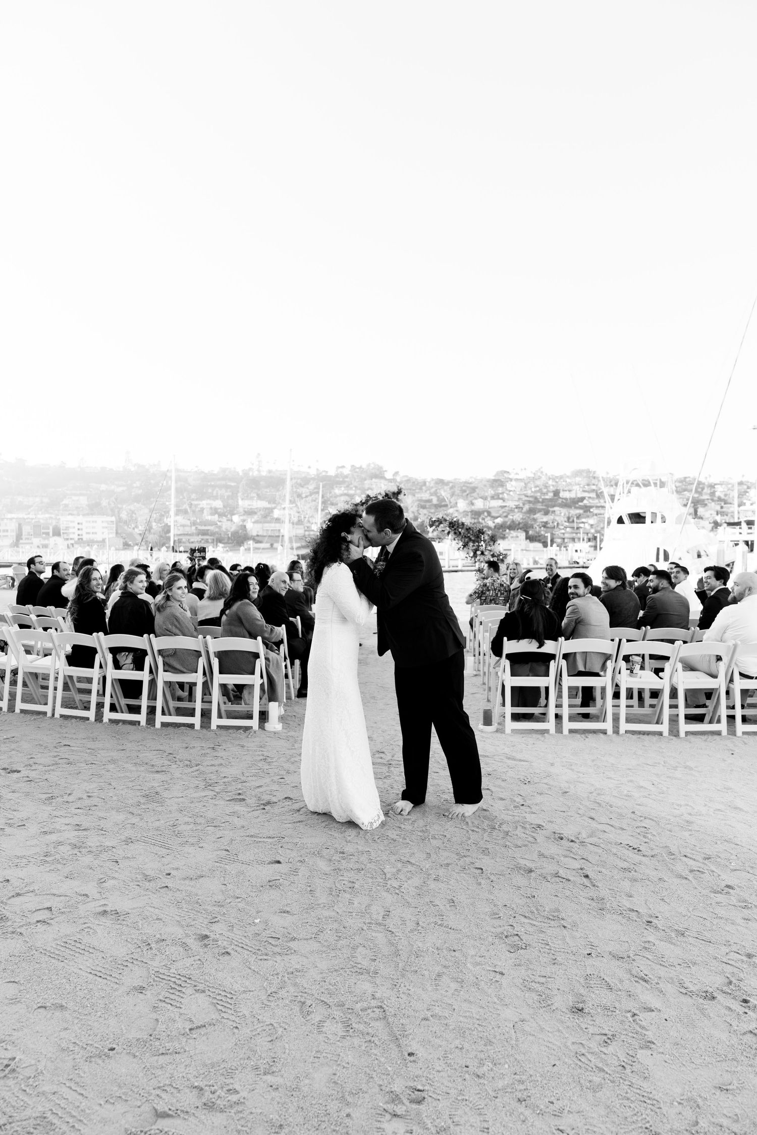 A couple shares their first kiss during a beach wedding ceremony with guests seated in white chairs behind them.