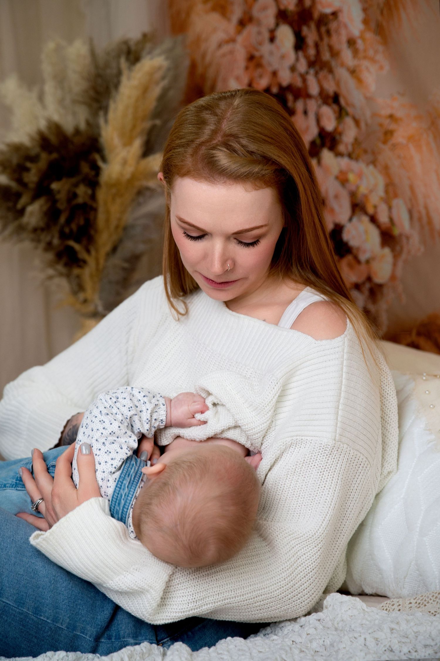 A tender moment as a mother in a white sweater nurses her baby in a cozy setting with dried flowers in the background.