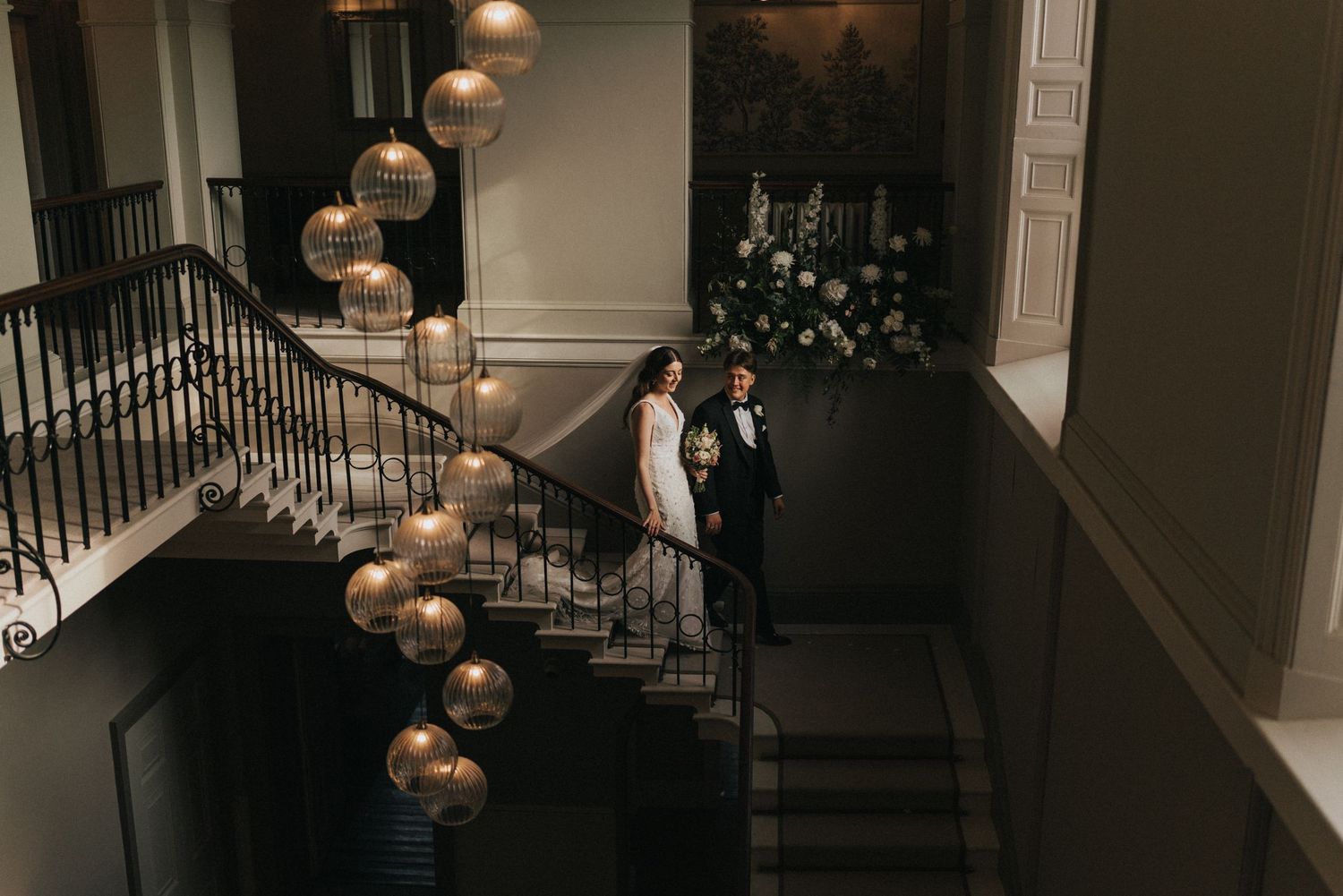A couple stands on an elegant staircase with cascading gold orb pendant lights at Saltmarshe Hall - taken by Grimsby photographer.