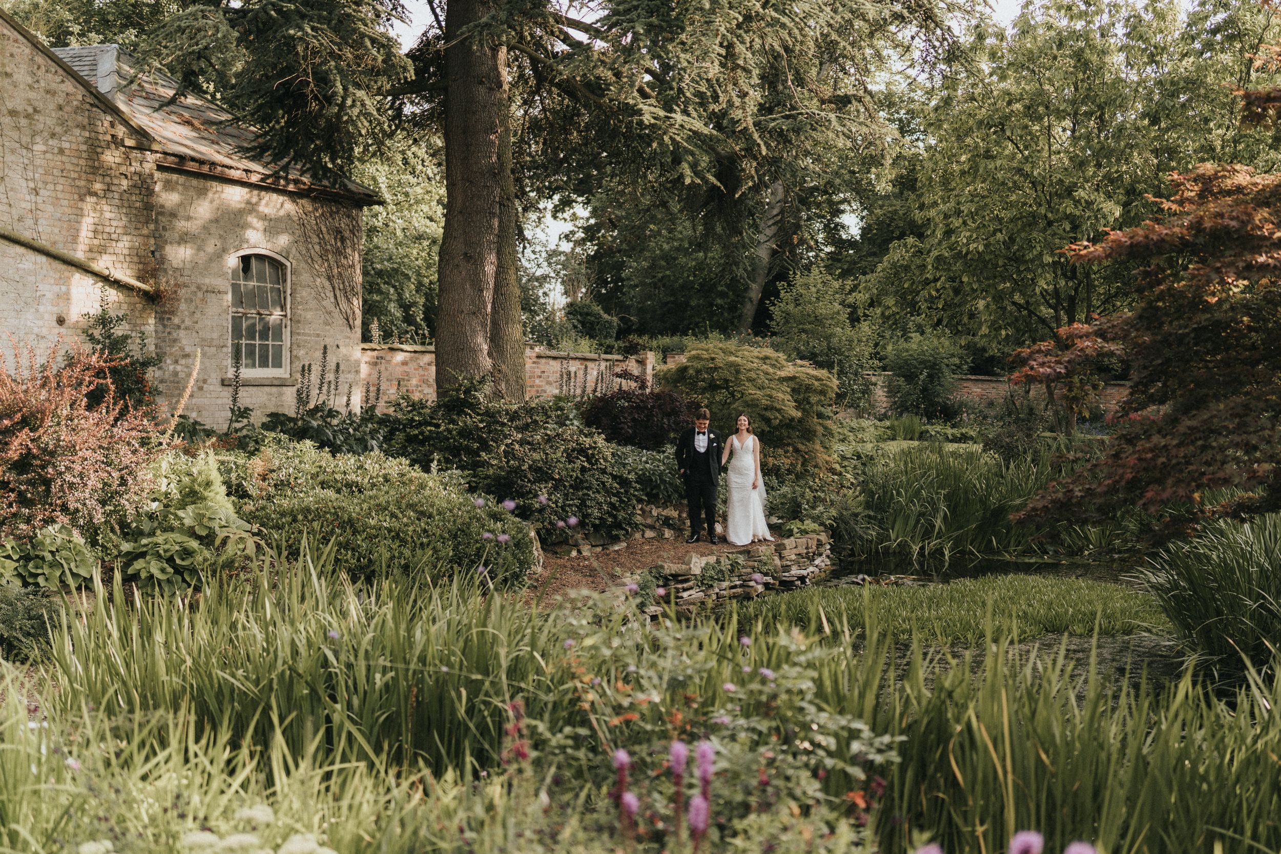 A wedding couple stands in a lush garden path surrounded by tall grasses and flowering plants at Saltmarshe Hall.