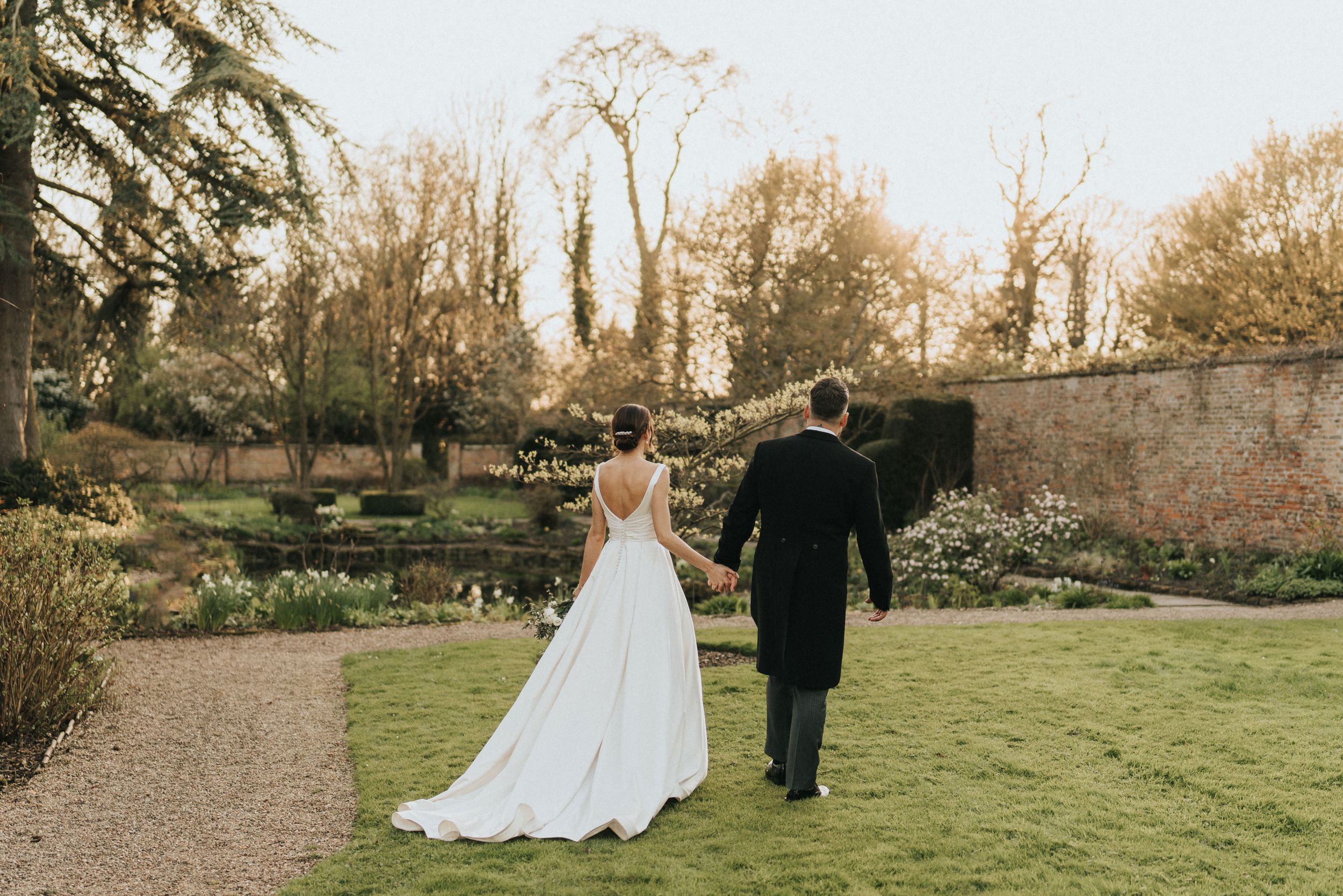Couple in formal wedding attire walking through a walled garden at sunset with daffodils blooming at Saltmarshe Hall.
