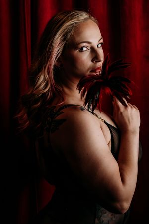 Dramatic portrait against red curtain backdrop featuring blonde hair and black feathers in moody lighting. Boudoir Photography Michigan