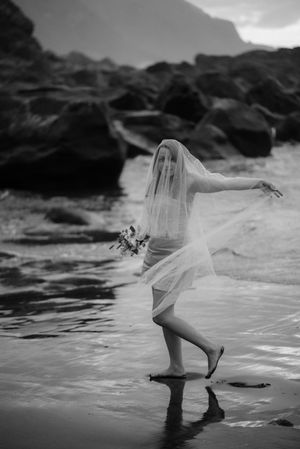 Black and white photograph of a flowing dress caught in ocean waves against rocky coastline. Playa de Benijo. Love Story in Tenerife.