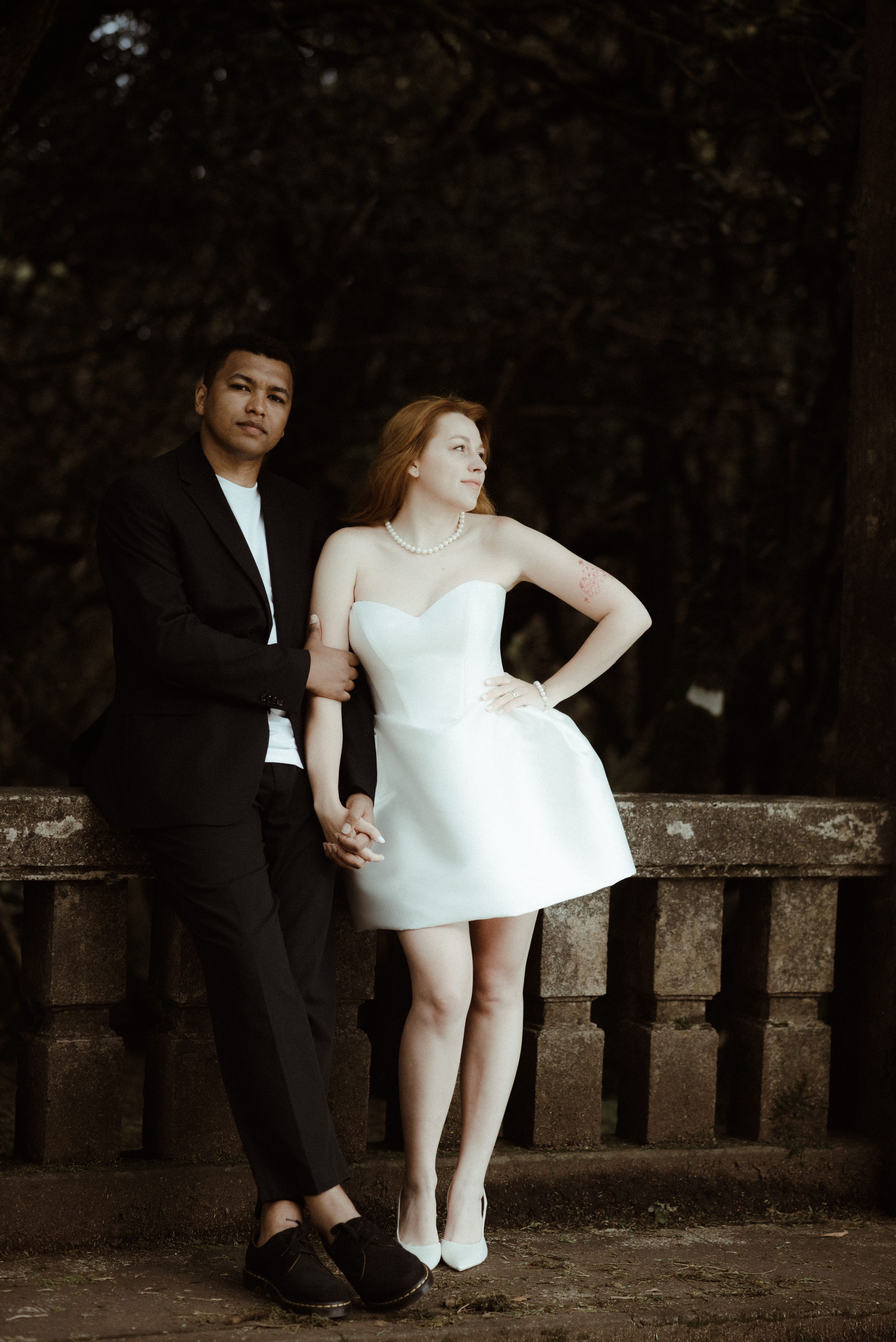 A romantic couple in formal attire sits together on an old stone wall in a dark wooded setting.