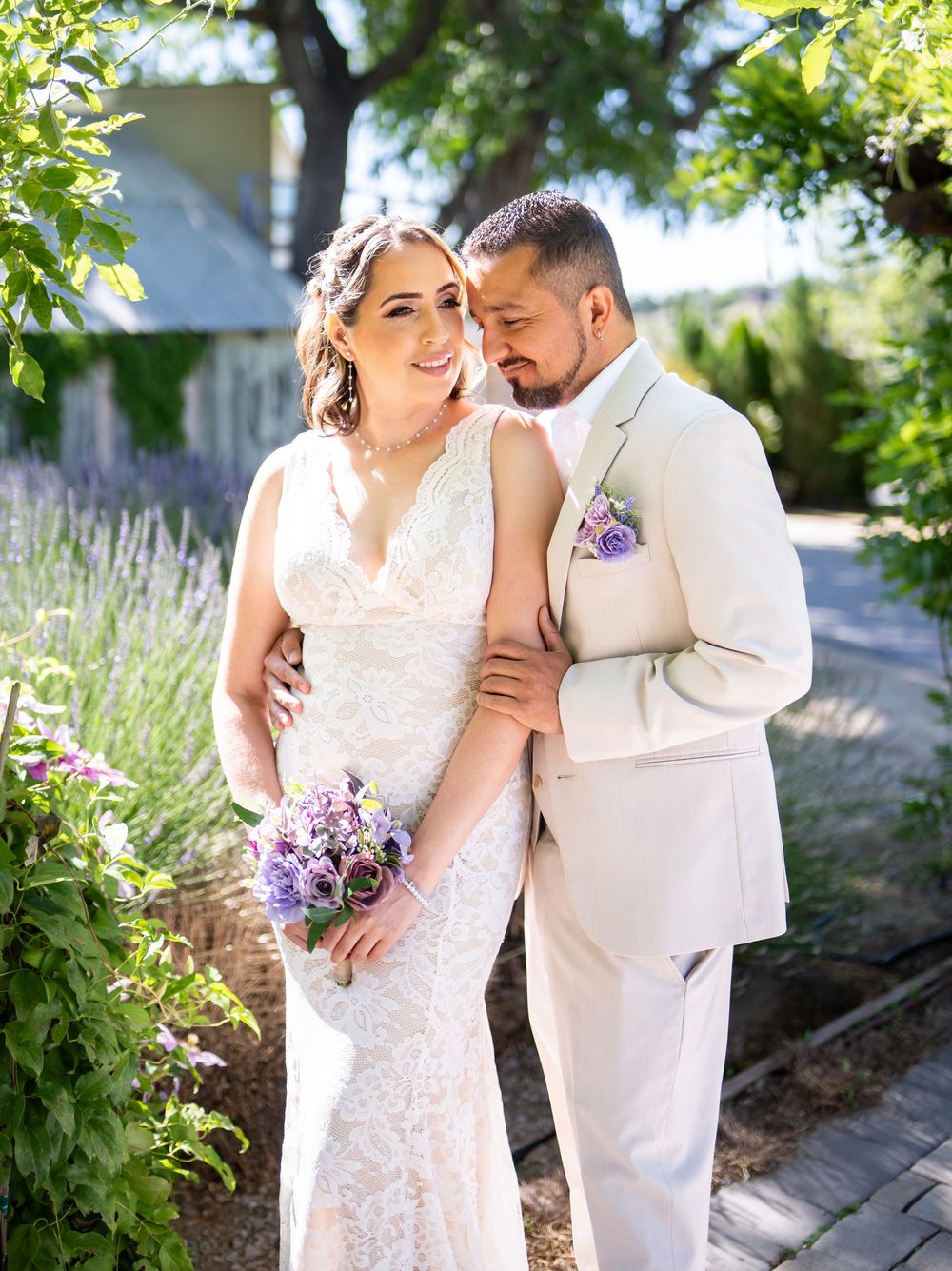 A couple shares an intimate moment among lavender and greenery in a sunlit garden on their wedding day.