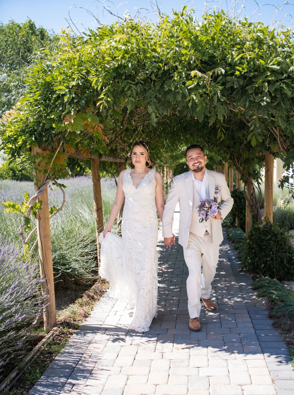 Couple in light-colored wedding attire walking and laughing under a lush green vine-covered garden archway during outdoor ceremony.