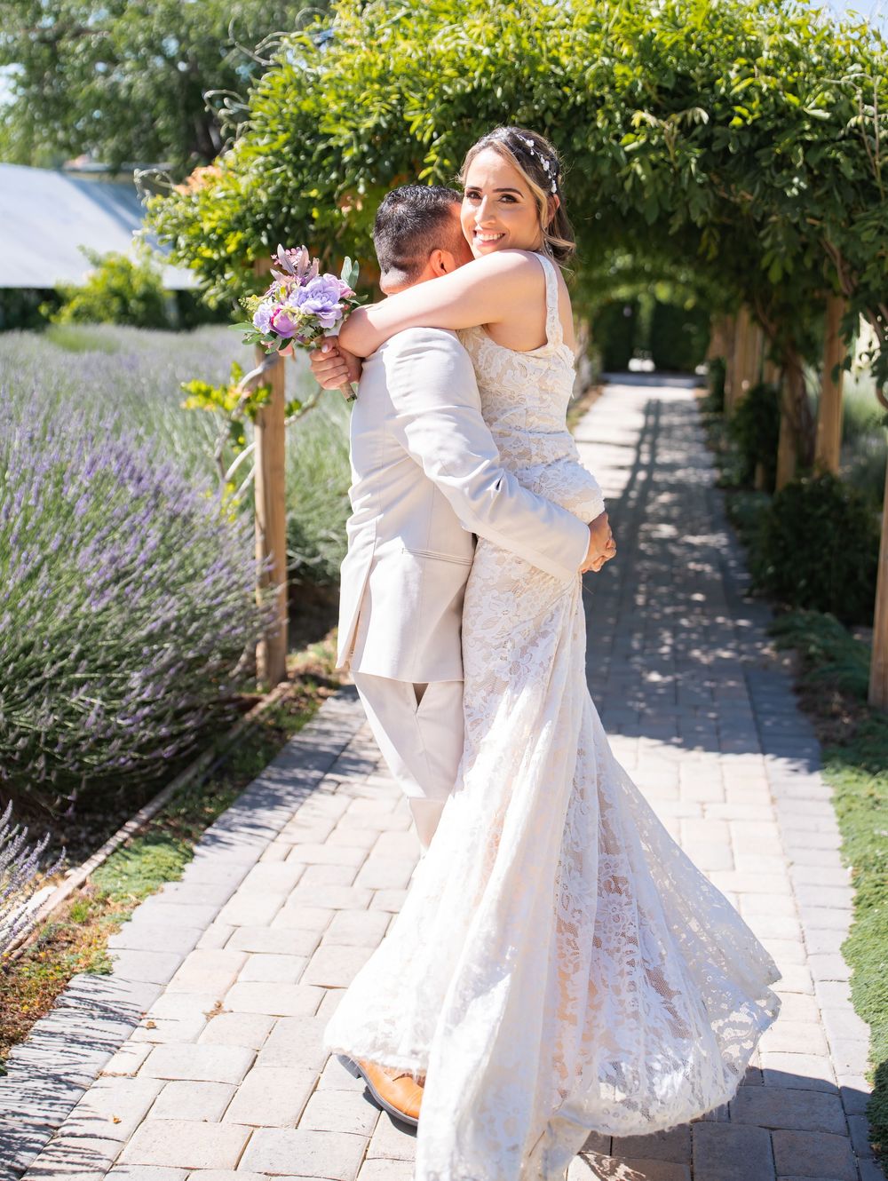 A couple embraces in an outdoor garden setting with lavender plants and a stone pathway on their wedding day.