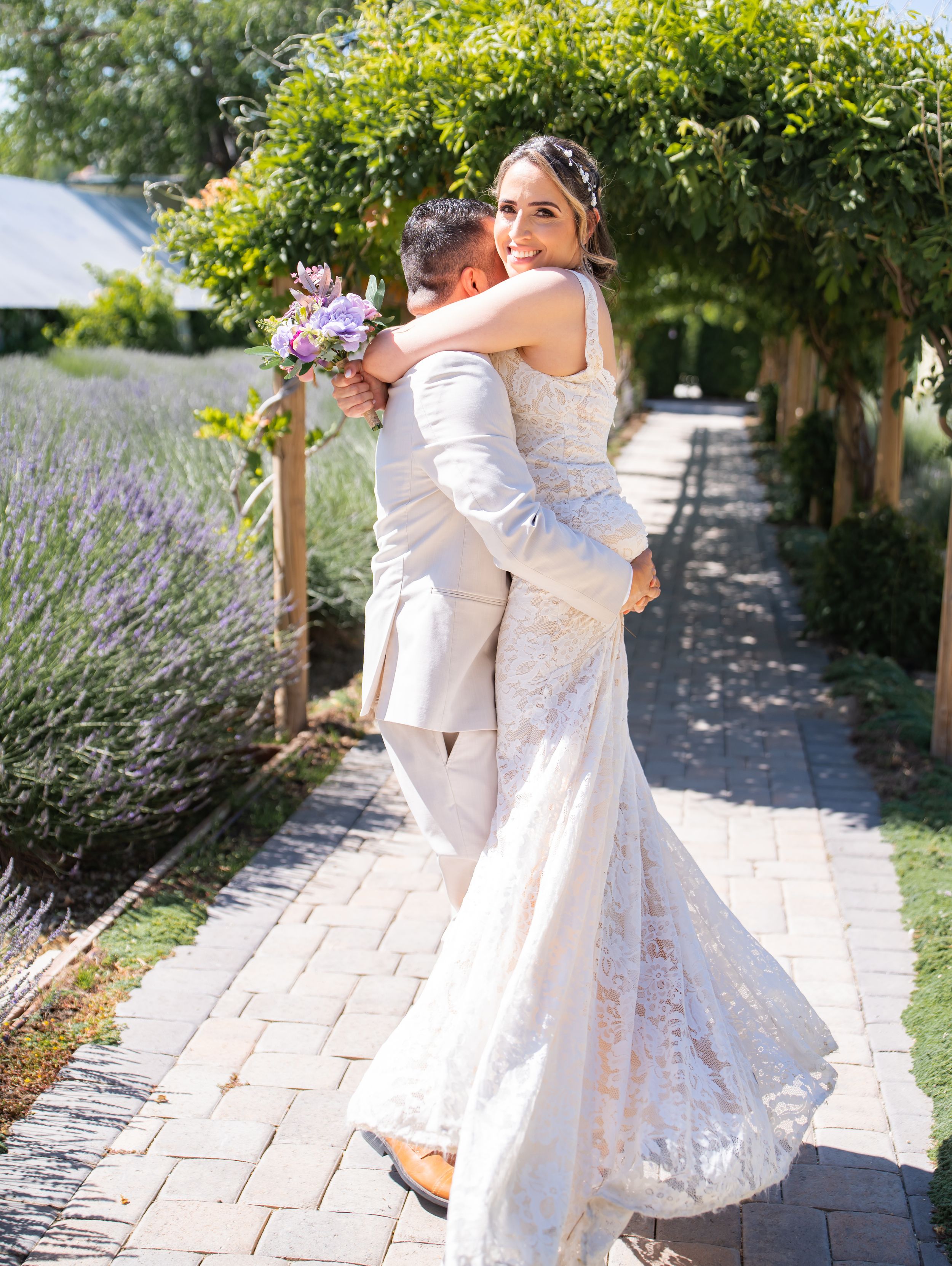A couple embraces in an outdoor garden setting with lavender plants and a stone pathway on their wedding day.