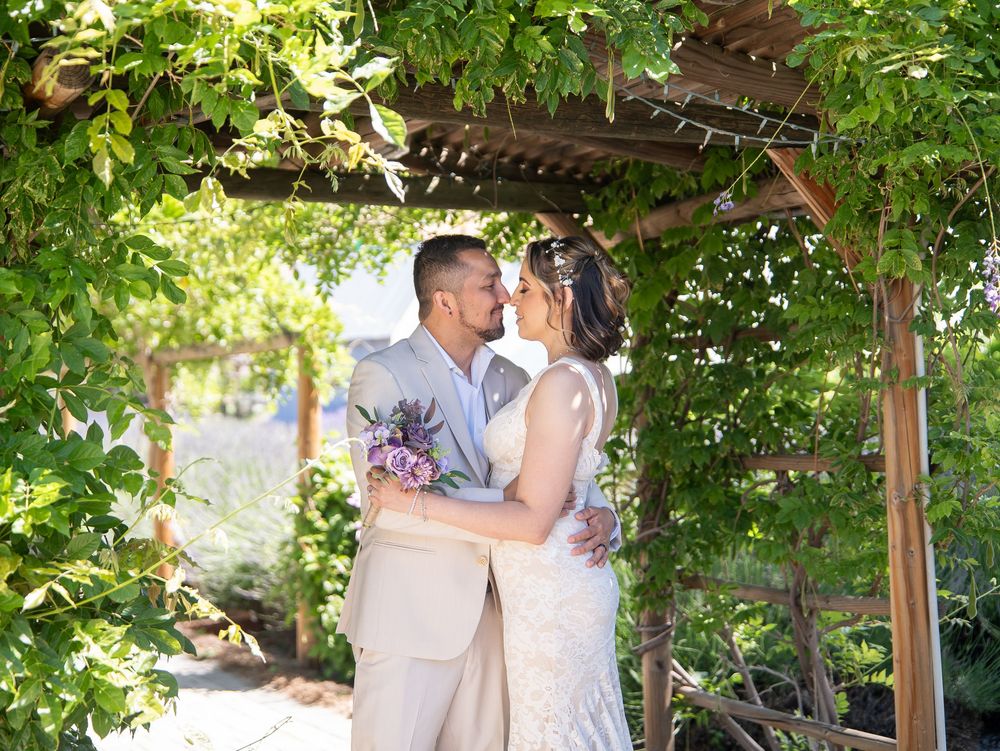 A couple shares a romantic moment under a lush garden pergola with climbing vines and dappled sunlight.