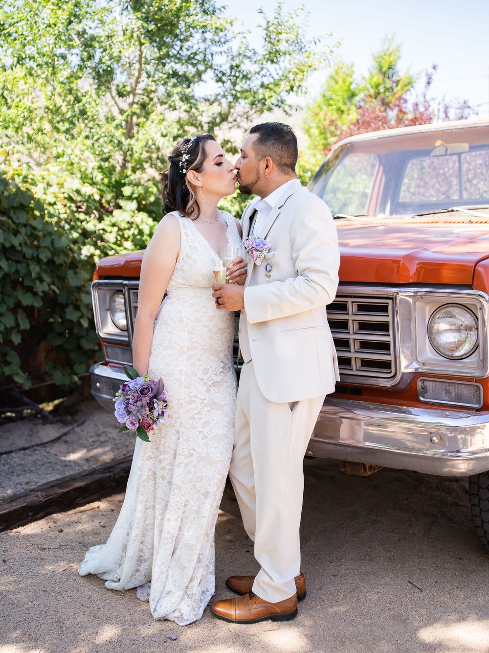 A couple in wedding attire shares romantic moments by a vintage orange truck on a sunny outdoor backdrop.