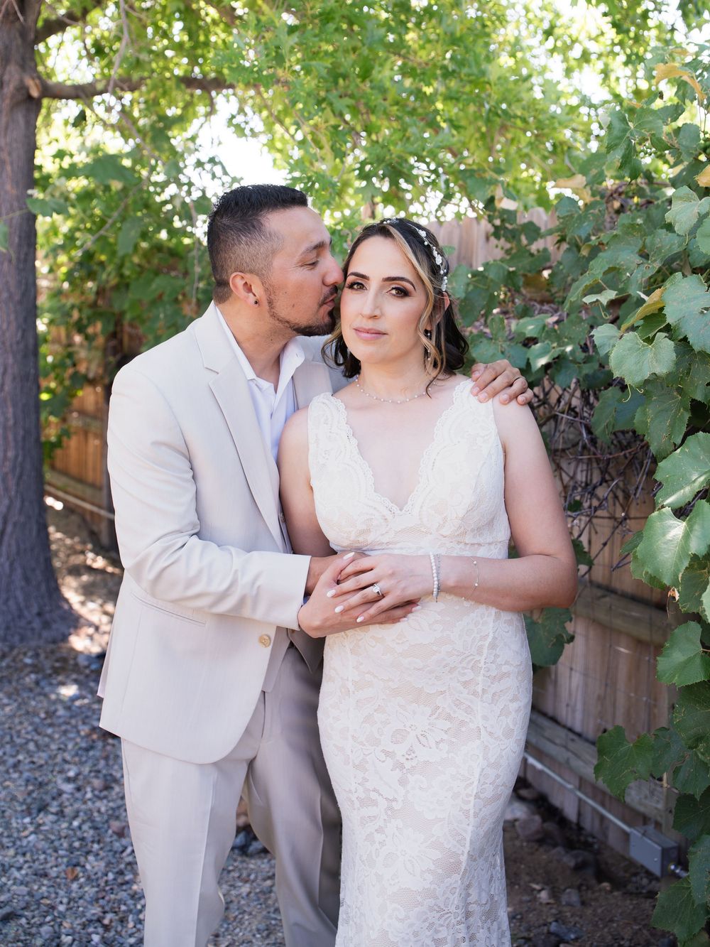 A couple stands together outdoors in their wedding attire against lush green foliage.