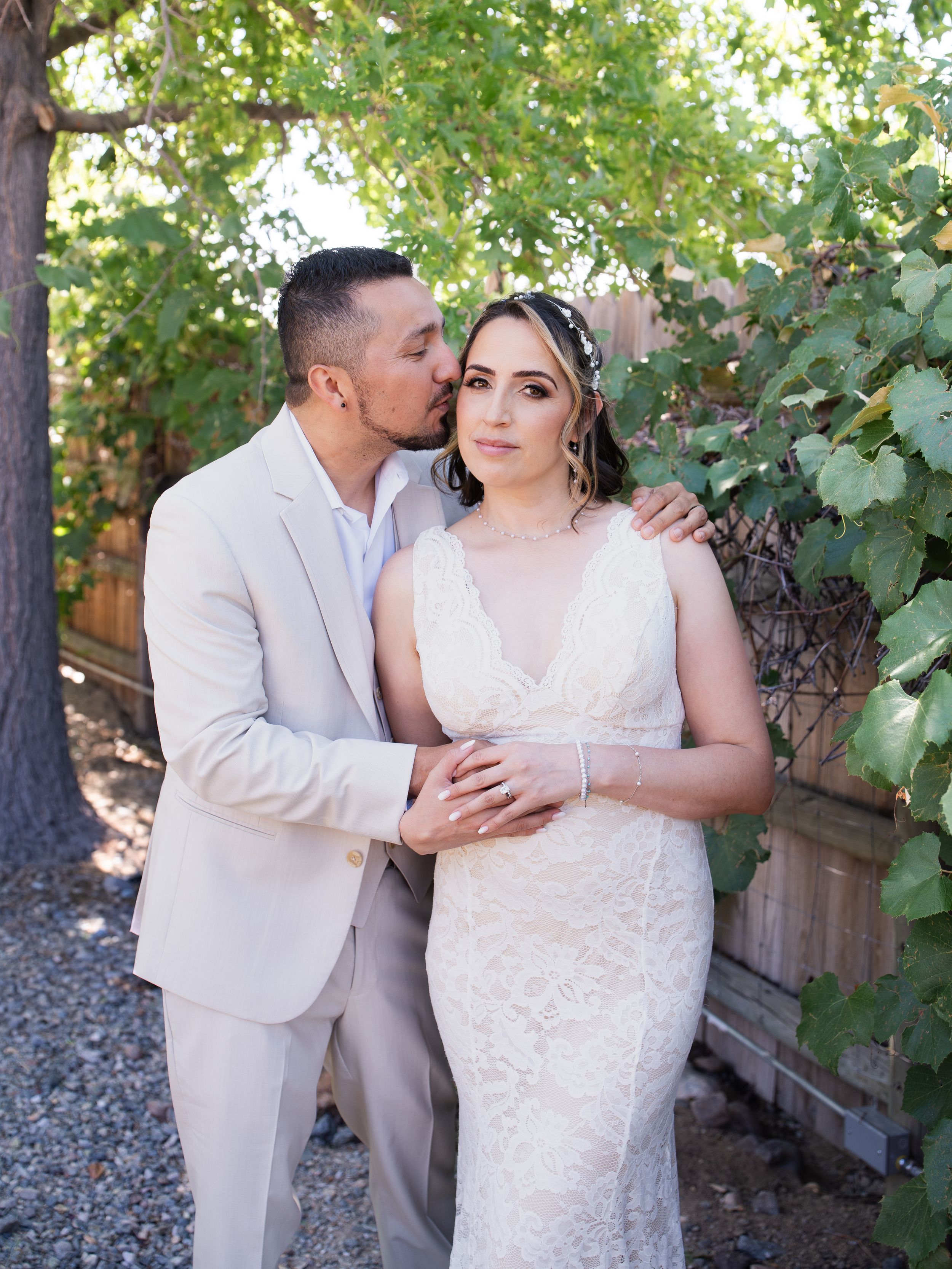 A couple stands together outdoors in their wedding attire against lush green foliage.